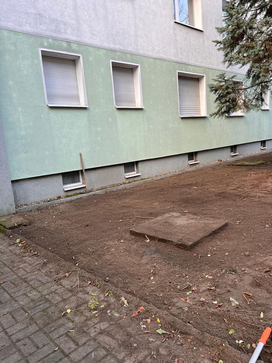 Vorbereiteter Erdstreifen an einer Hausfassade mit einer ausgerollten Rasensode, kurz vor der Rasenverlegung
