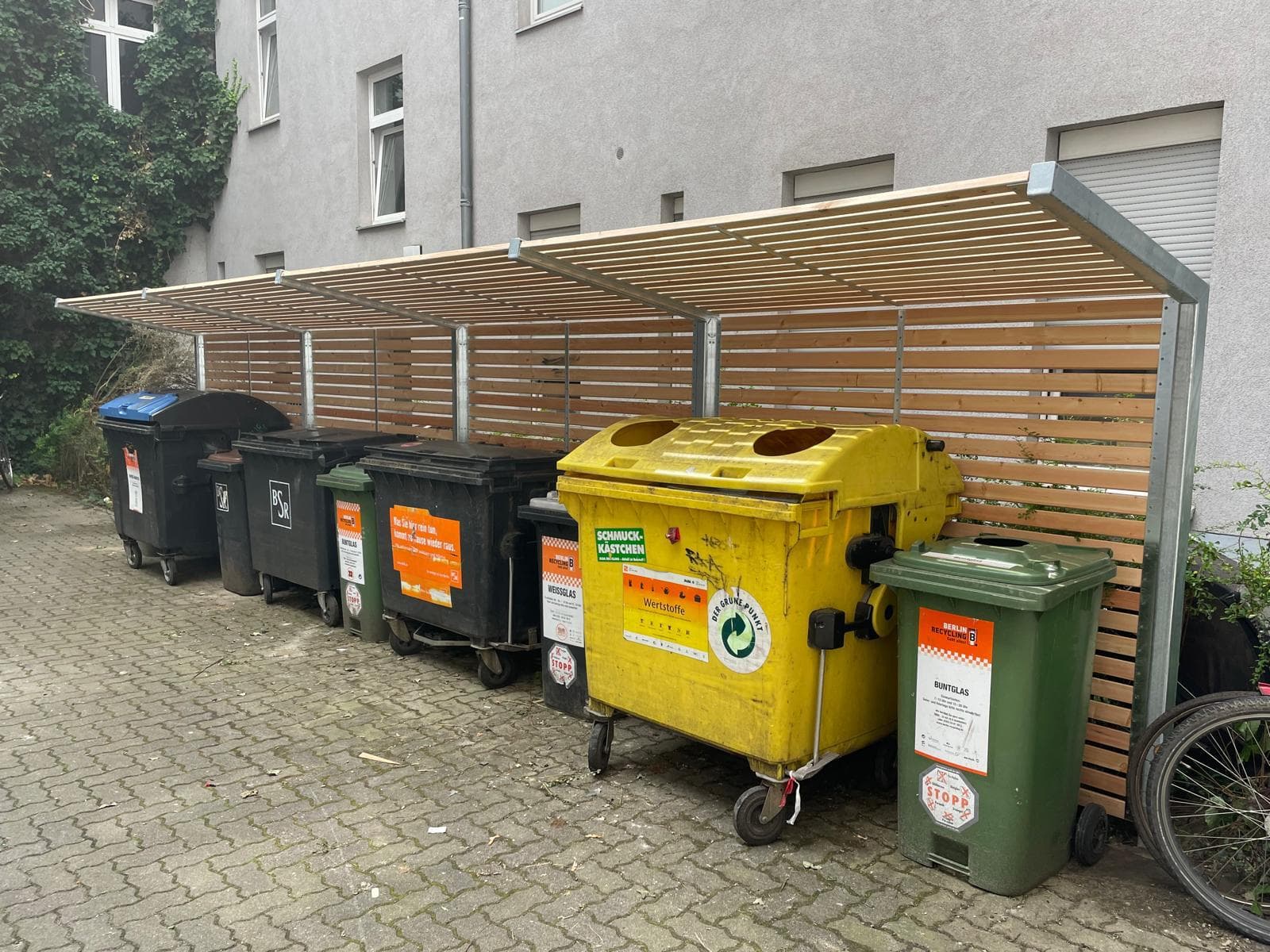 Seitenansicht einer von Sonne und Grün montierten Müllplatz-Einhausung mit Holzlatten-Sichtschutz in Berlin