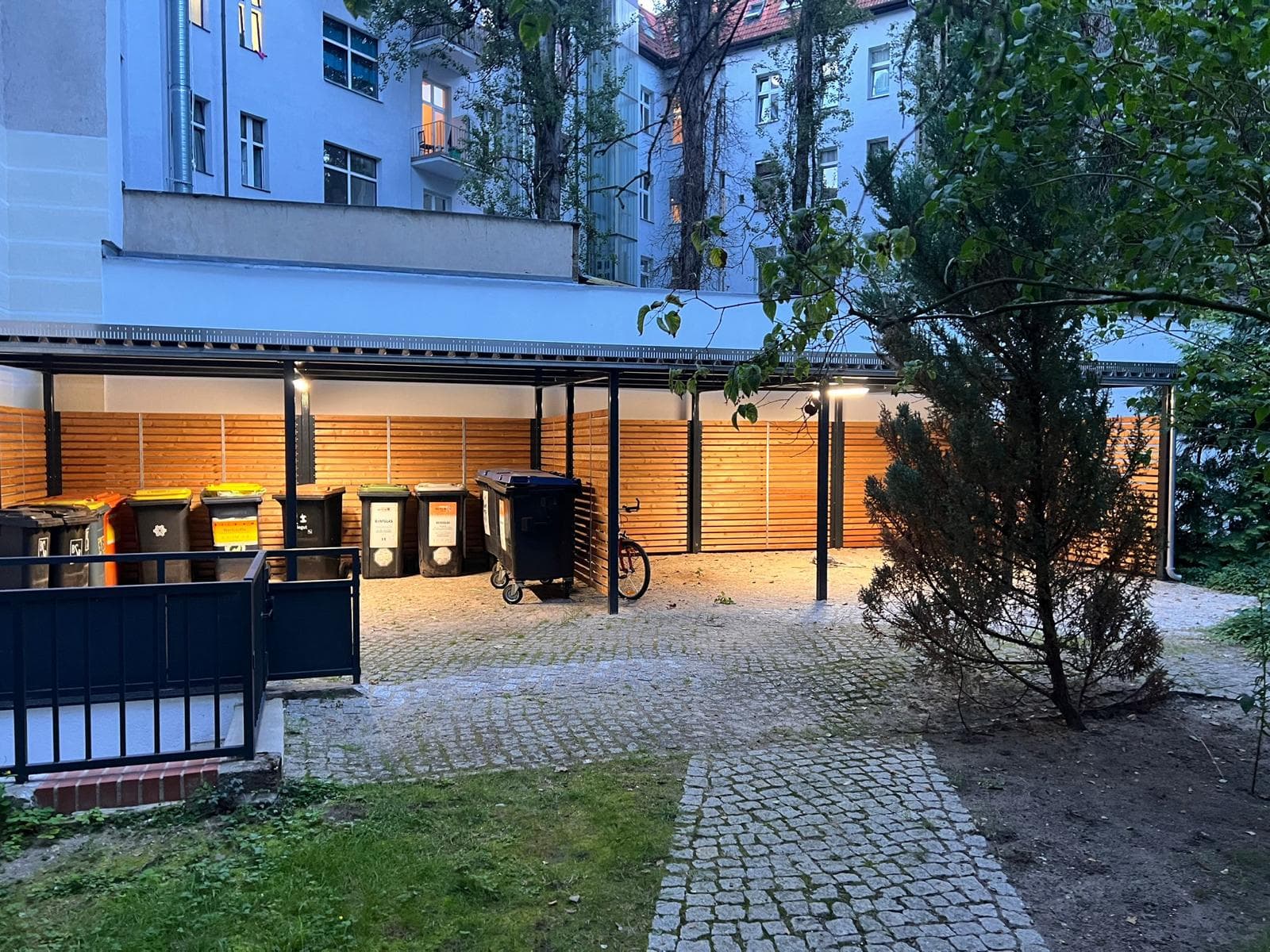 Beleuchtete Müllplatz-Überdachung mit Holzlatten-Sichtschutz in einem begrünten Berliner Innenhof bei Dämmerung